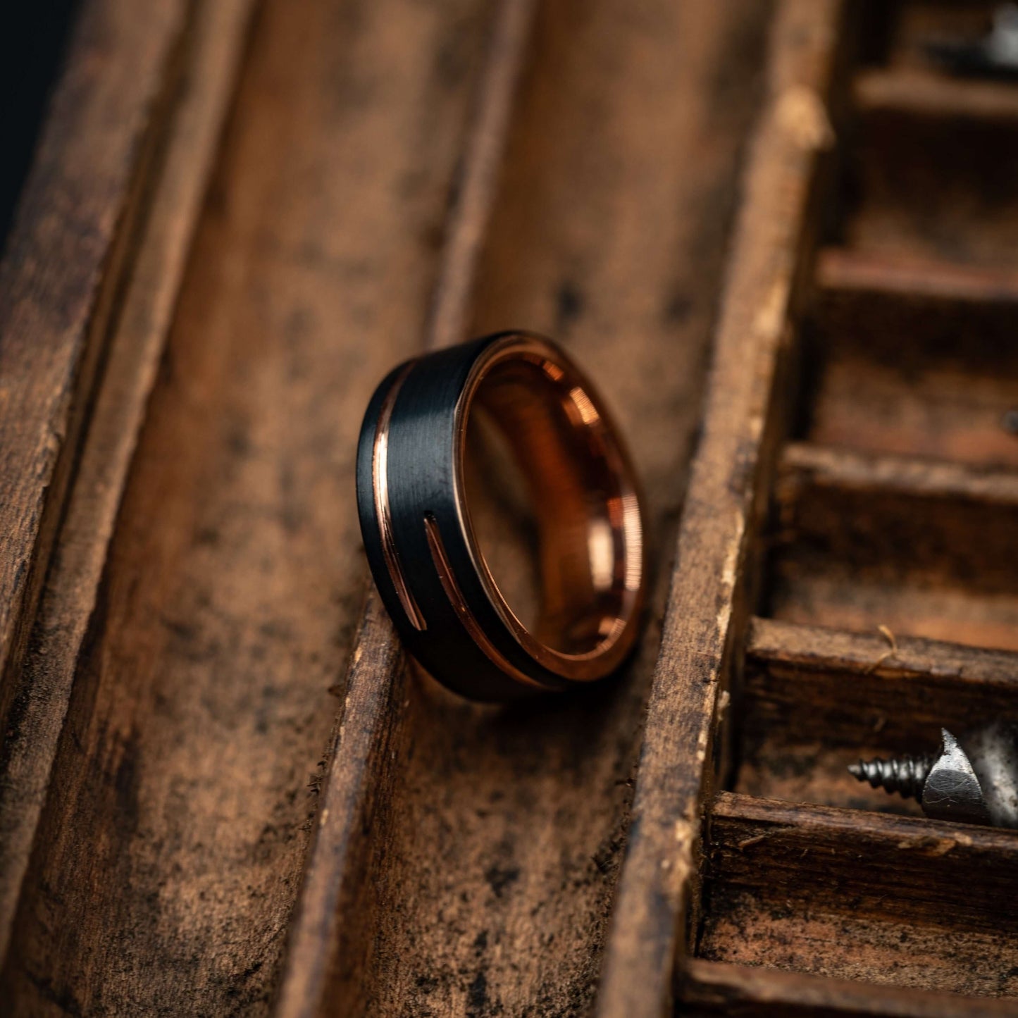 Black and rose gold 8mm wedding band resting in a wooden tray with screws, showcasing its unique design.