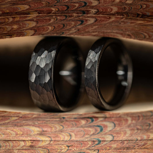 Black hammered tungsten wedding rings set for couples, showcasing their unique texture and durability against a wood backdrop.