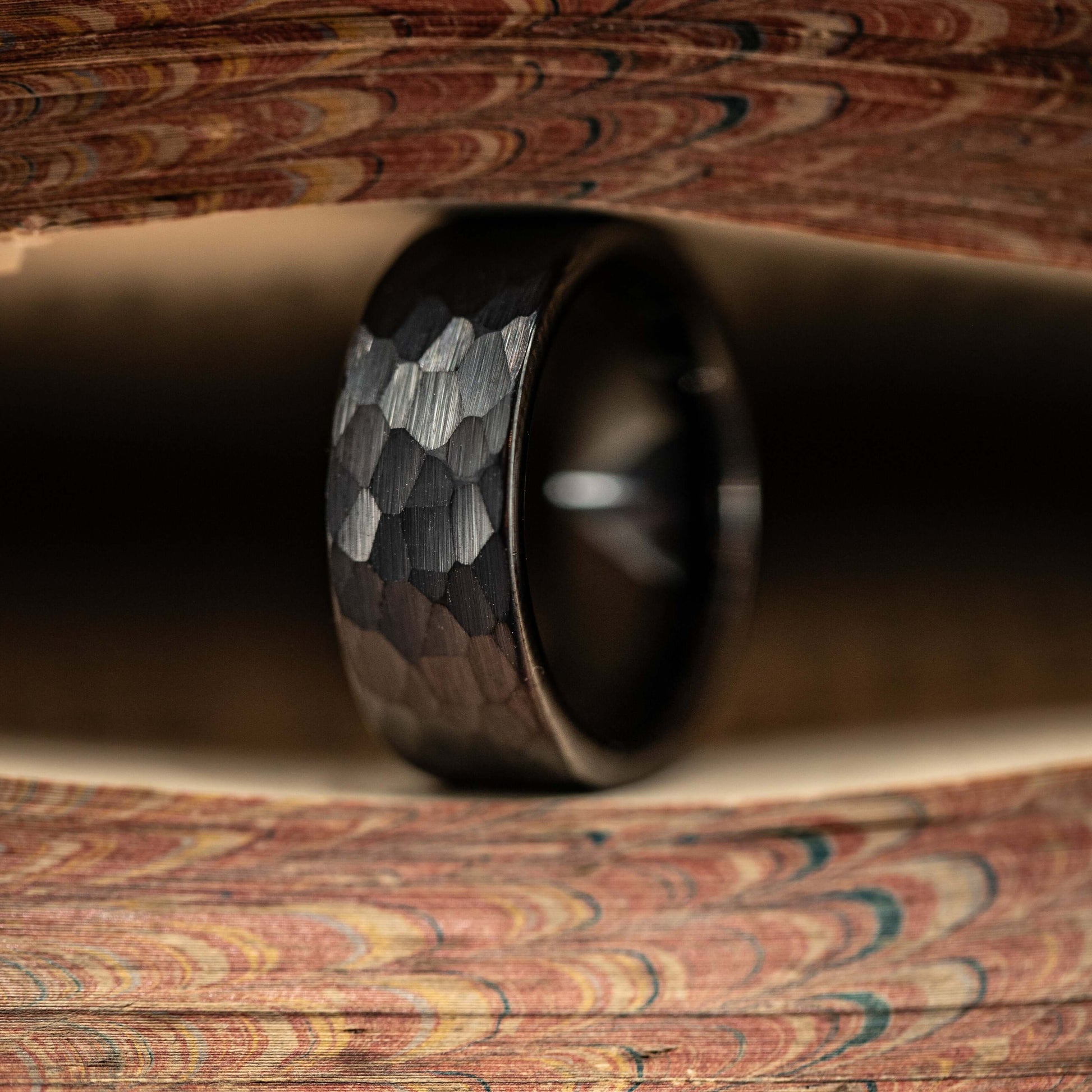 Hammered black tungsten wedding ring displayed between two wooden surfaces, showcasing its unique texture and finish.