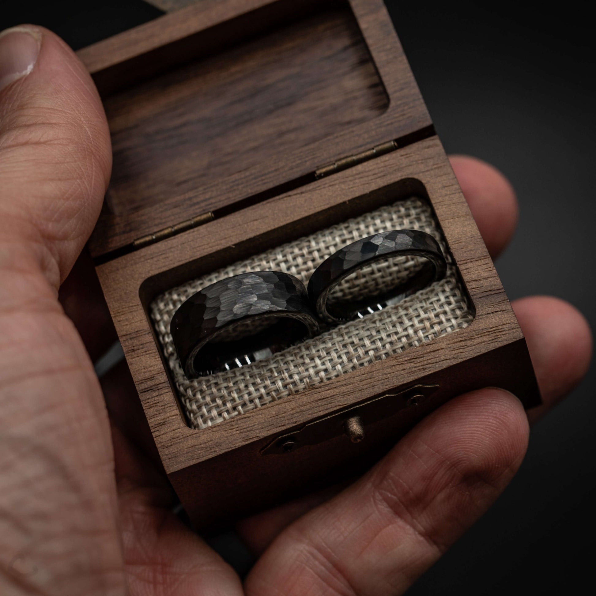 Black hammered tungsten wedding rings displayed in a wooden box, showcasing their bold and rustic design.
