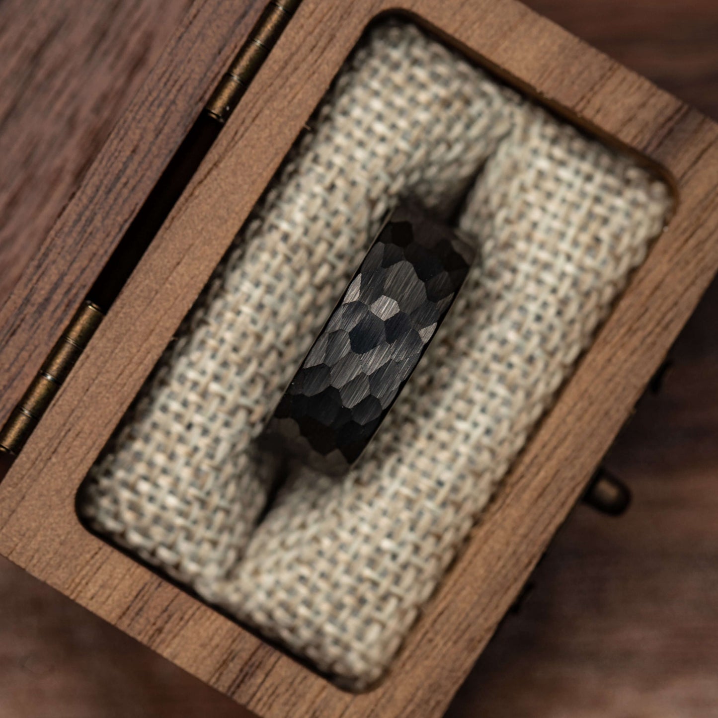 Hammered black tungsten wedding band displayed in a wooden box with burlap padding. Unique men's engagement ring.