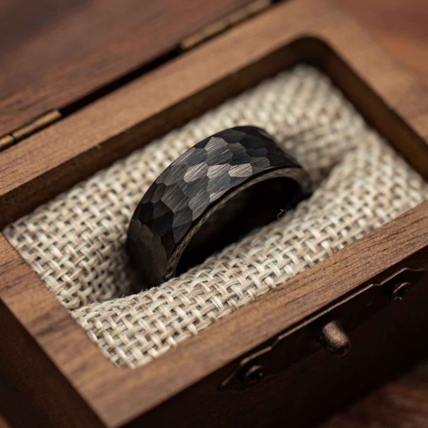 Hammered black tungsten wedding ring displayed in a wooden box lined with fabric, showcasing its bold design.