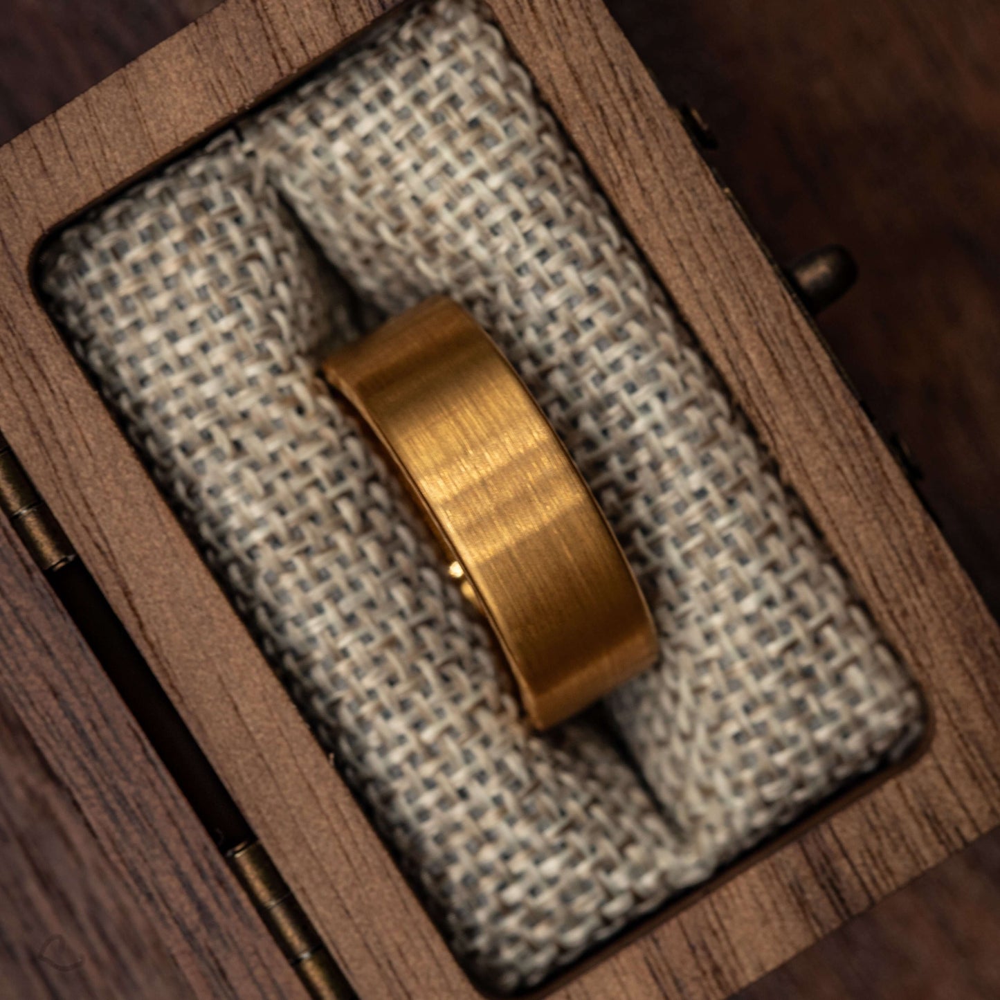 Gold brushed tungsten wedding band displayed on textured fabric in a wooden box.