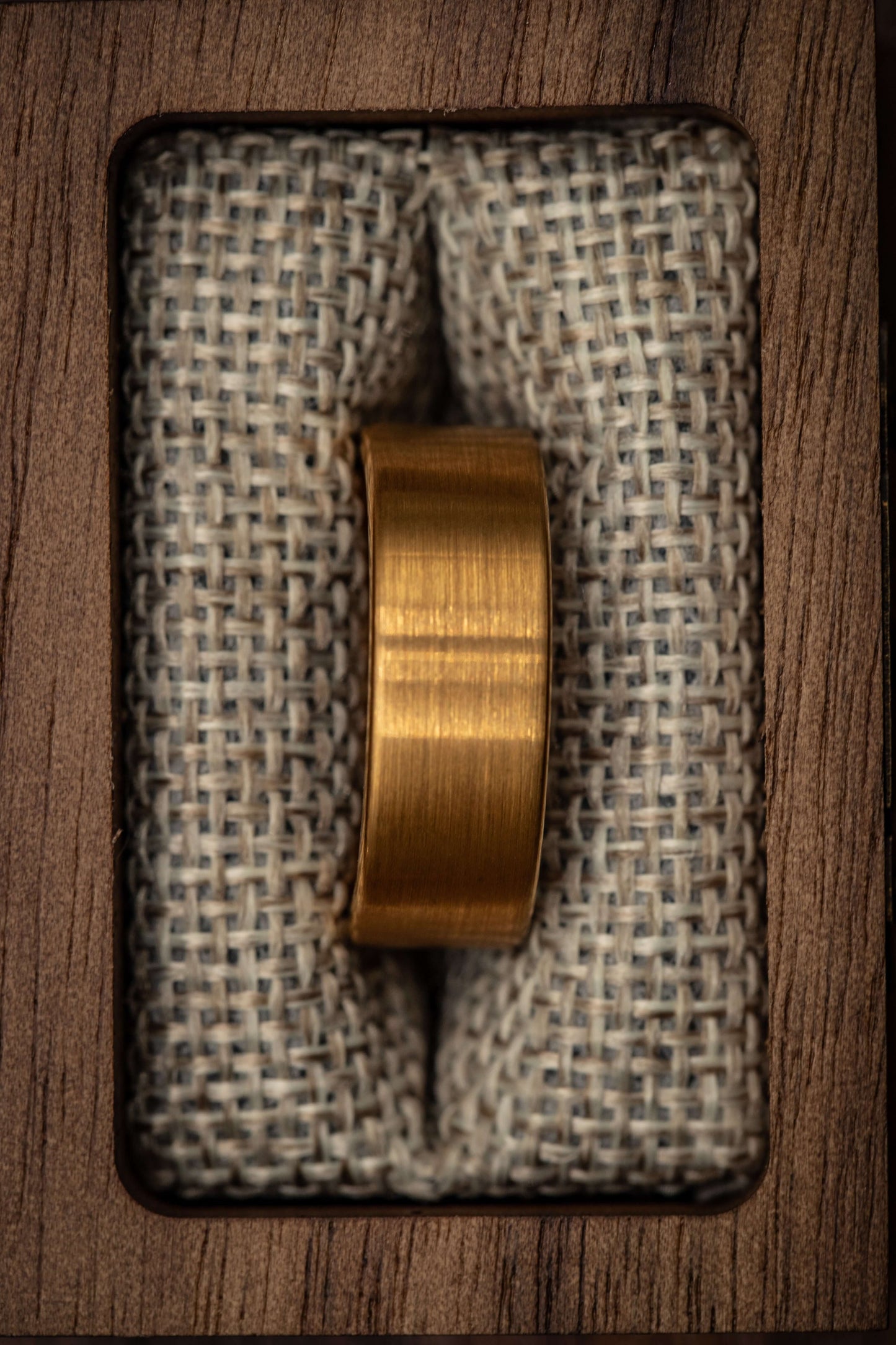 Gold brushed tungsten wedding ring displayed on textured fabric in a wooden box.