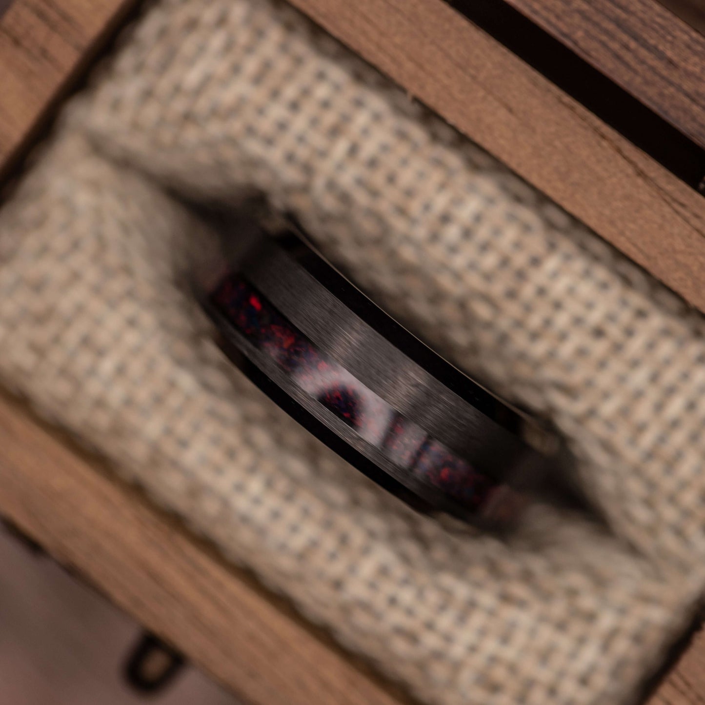 Black brushed tungsten wedding band with black fire opal, nestled in natural fabric within a wooden box.