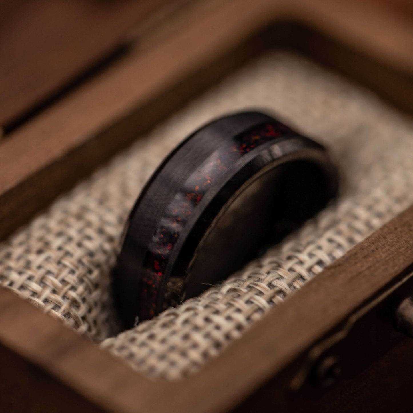 Black brushed tungsten ring with fire opal channel displayed in a wooden box on burlap fabric.