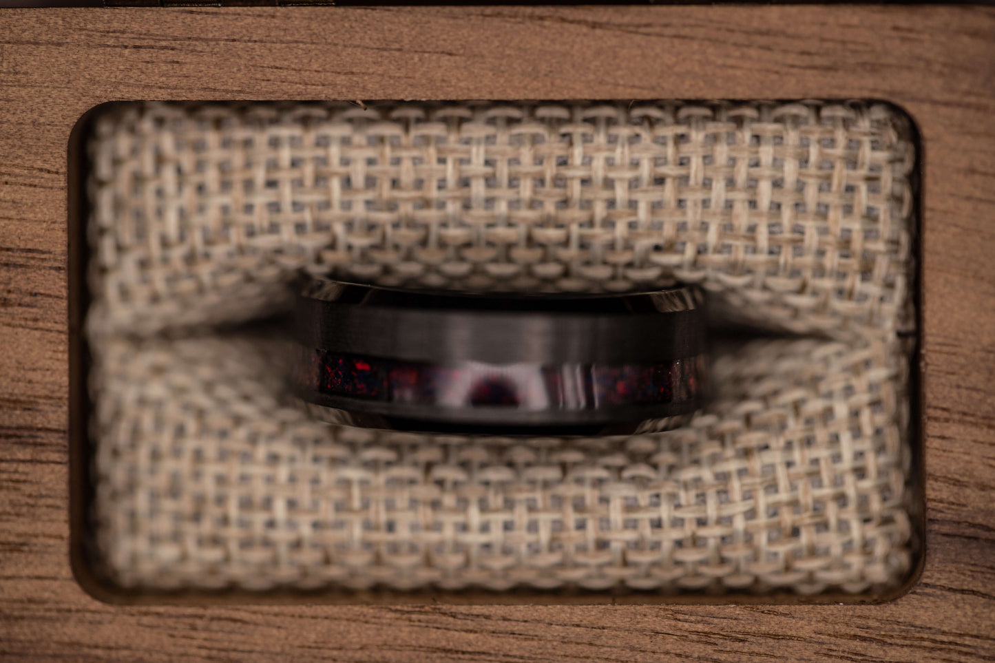 Black brushed tungsten ring featuring a channel of black fire opal, displayed in a wooden box with textured fabric.
