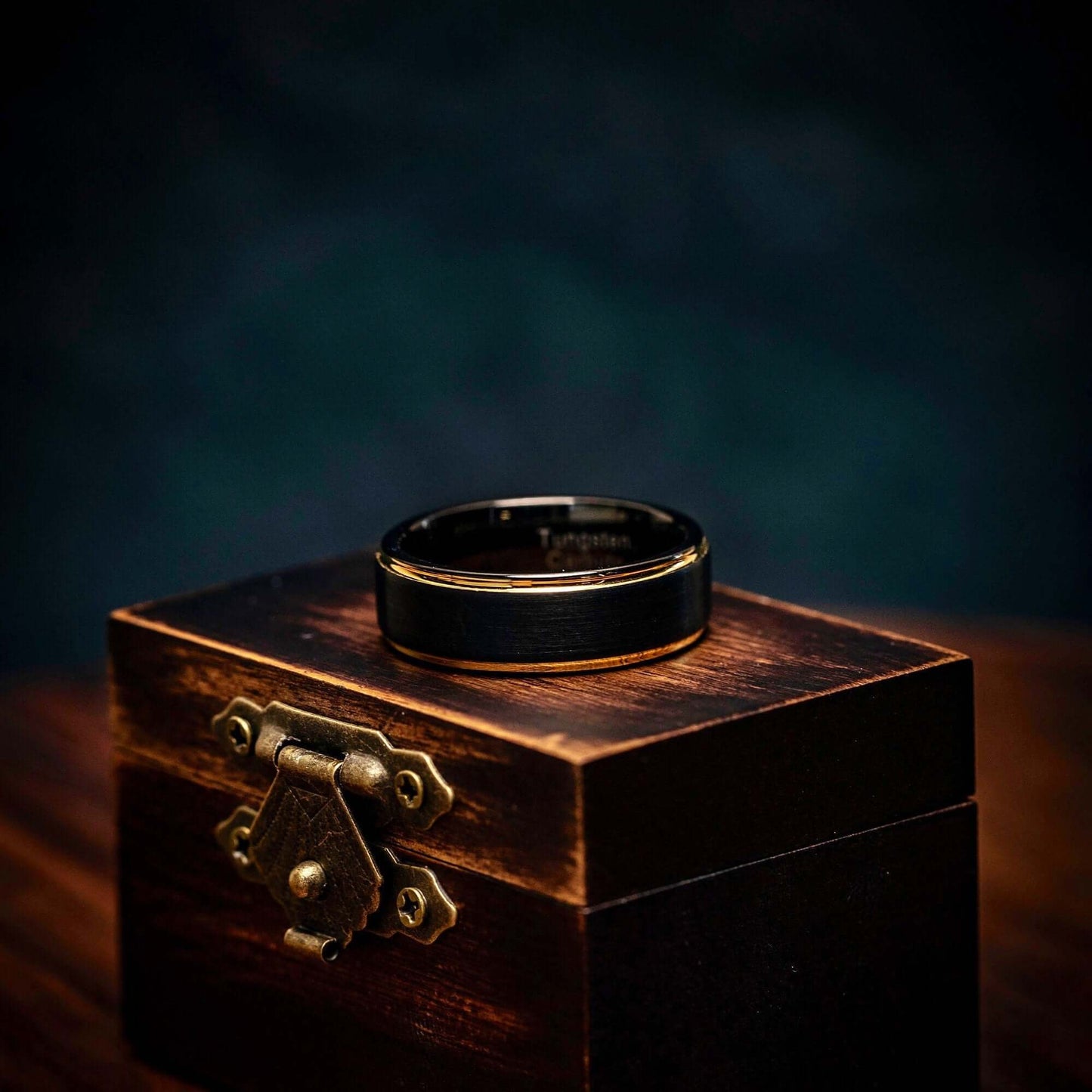 Black tungsten wedding ring with 18k Yellow Gold Step edges laying on its side on top of walnut ring box