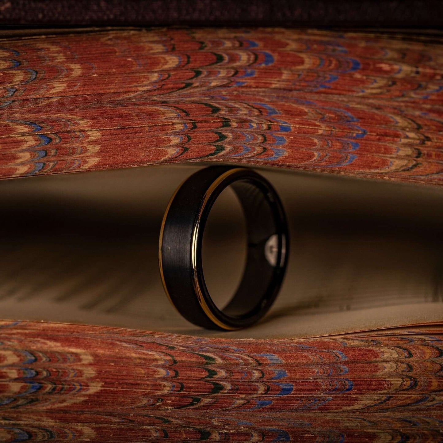 Black tungsten wedding ring with 18k Yellow Gold Step edges standing on its side between pages of a book
