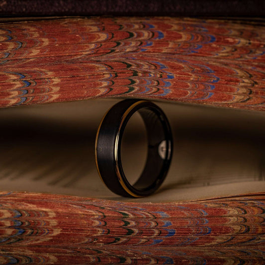 Black tungsten wedding ring with 18k Yellow Gold Step edges standing on its side between pages of a book