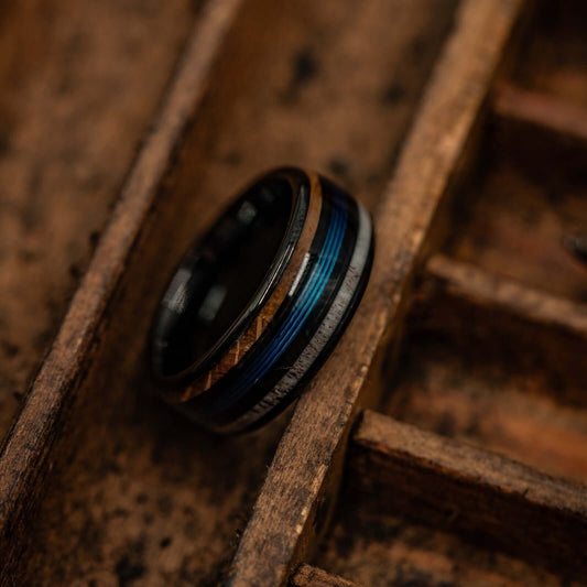 A men's 8mm black tungsten wedding band with antler, whiskey barrel wood and blue fishing line inlays, resting at an angle in a weathered wooden tray.