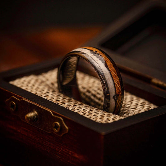 A men's 8mm tungsten wedding band with deer antler and whiskey barrel wood inlay, propped upright inside an open walnut ring box lined with burlap.