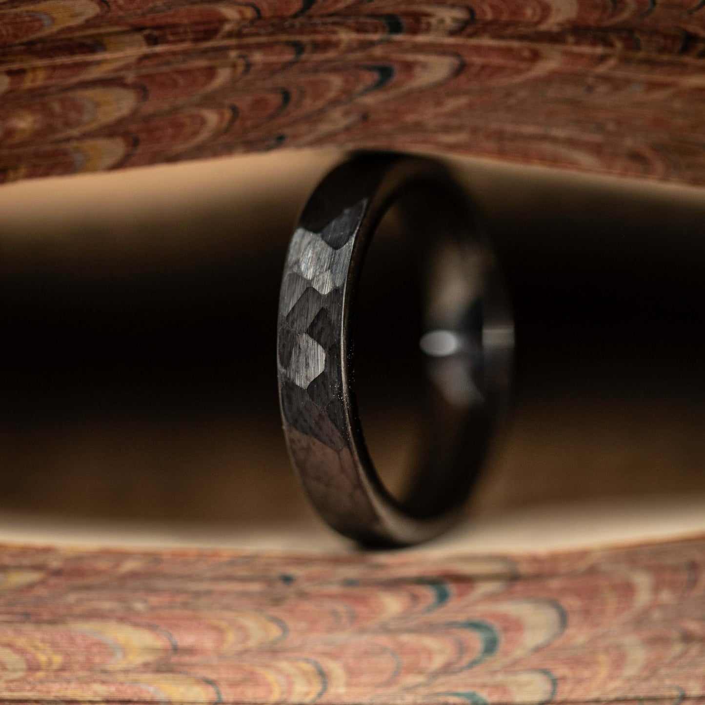 Black hammered tungsten wedding band showcased between colorful wood textures, highlighting its unique design and durability.