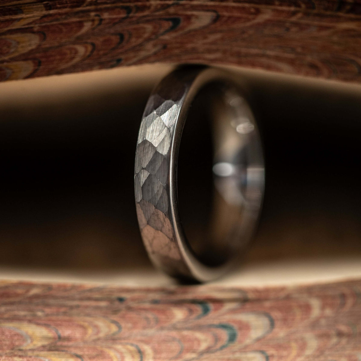 A close-up, centered shot of the Women’s 4mm Silver Hammered Tungsten Wedding Ring stands upright between the pages of an antique book with marbled, swirl-patterned edges. The soft lighting highlights the ring’s textured silver hammered surface, emphasizing its unique craftsmanship.
