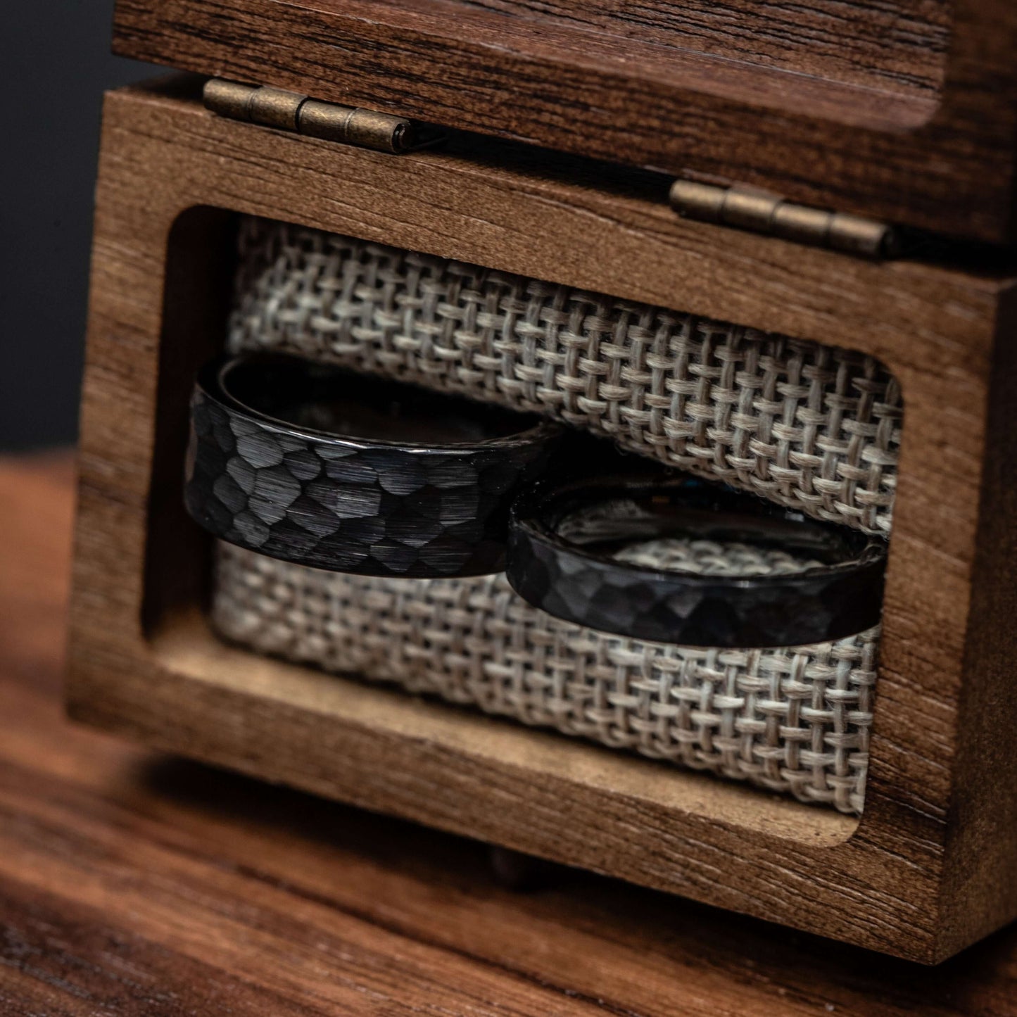 Two hammered black tungsten wedding rings displayed in a wooden box with fabric lining.