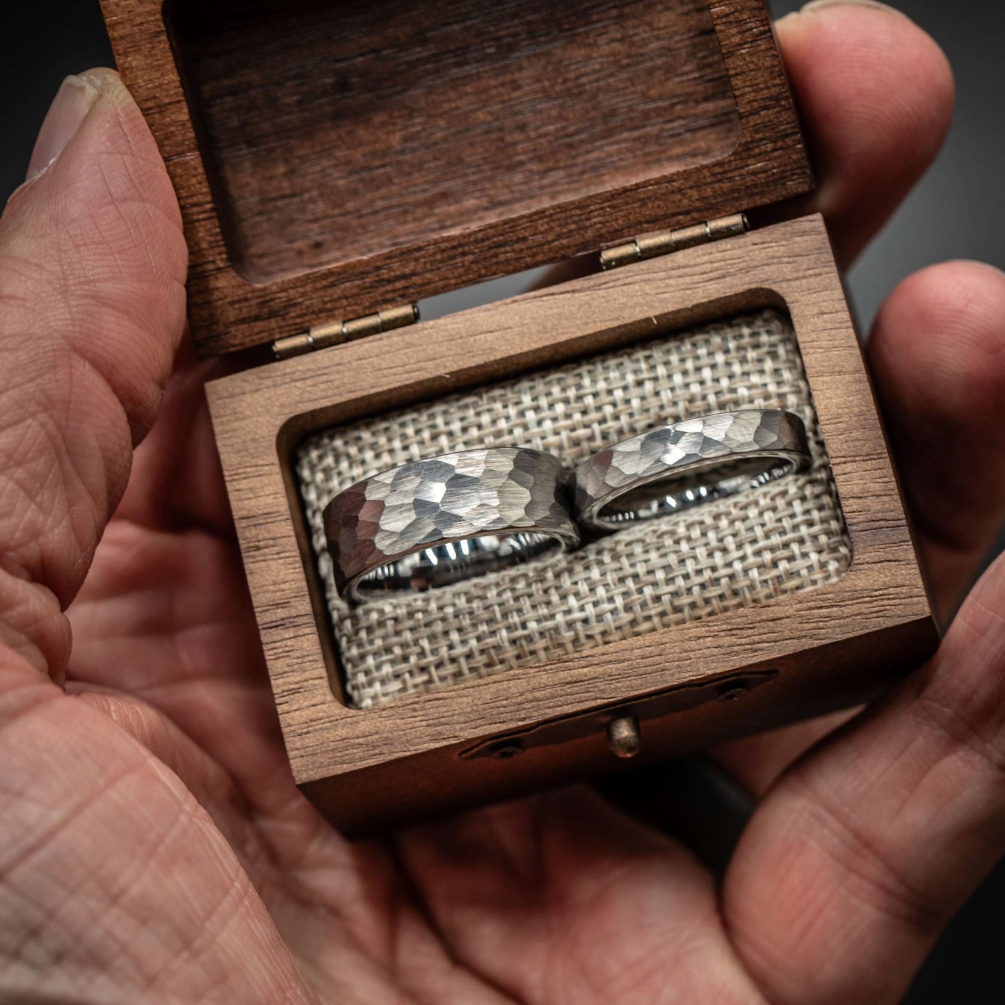 Hand holding a wooden box containing a pair of hammered silver tungsten wedding rings.