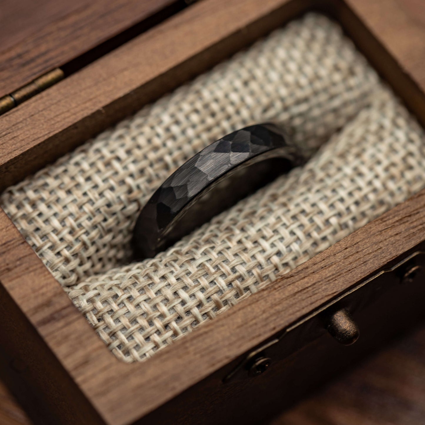 Hammered black tungsten wedding ring displayed on fabric in a wooden box. Unique and durable 4mm band.