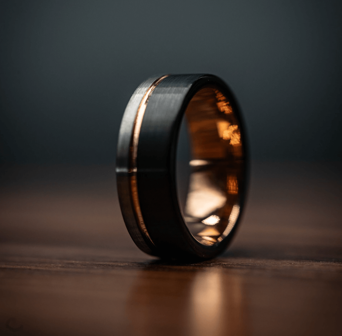 Black and silver wedding ring with rose gold inlay on wooden surface.