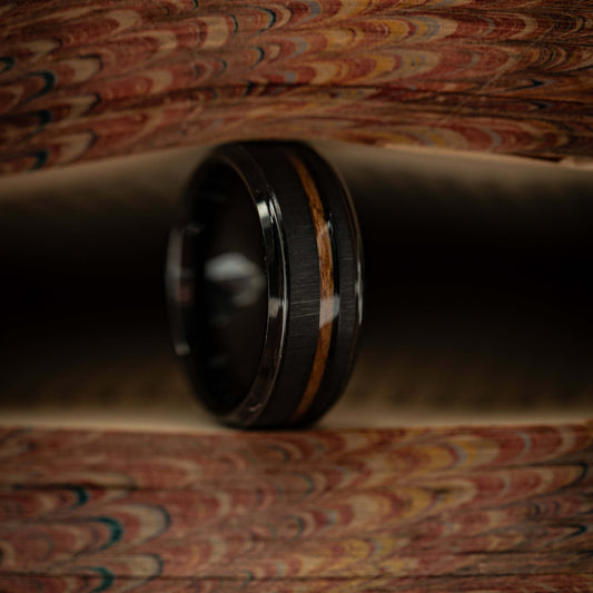 Black brushed tungsten and Hawaiian Koa wood wedding ring displayed on a textured surface.