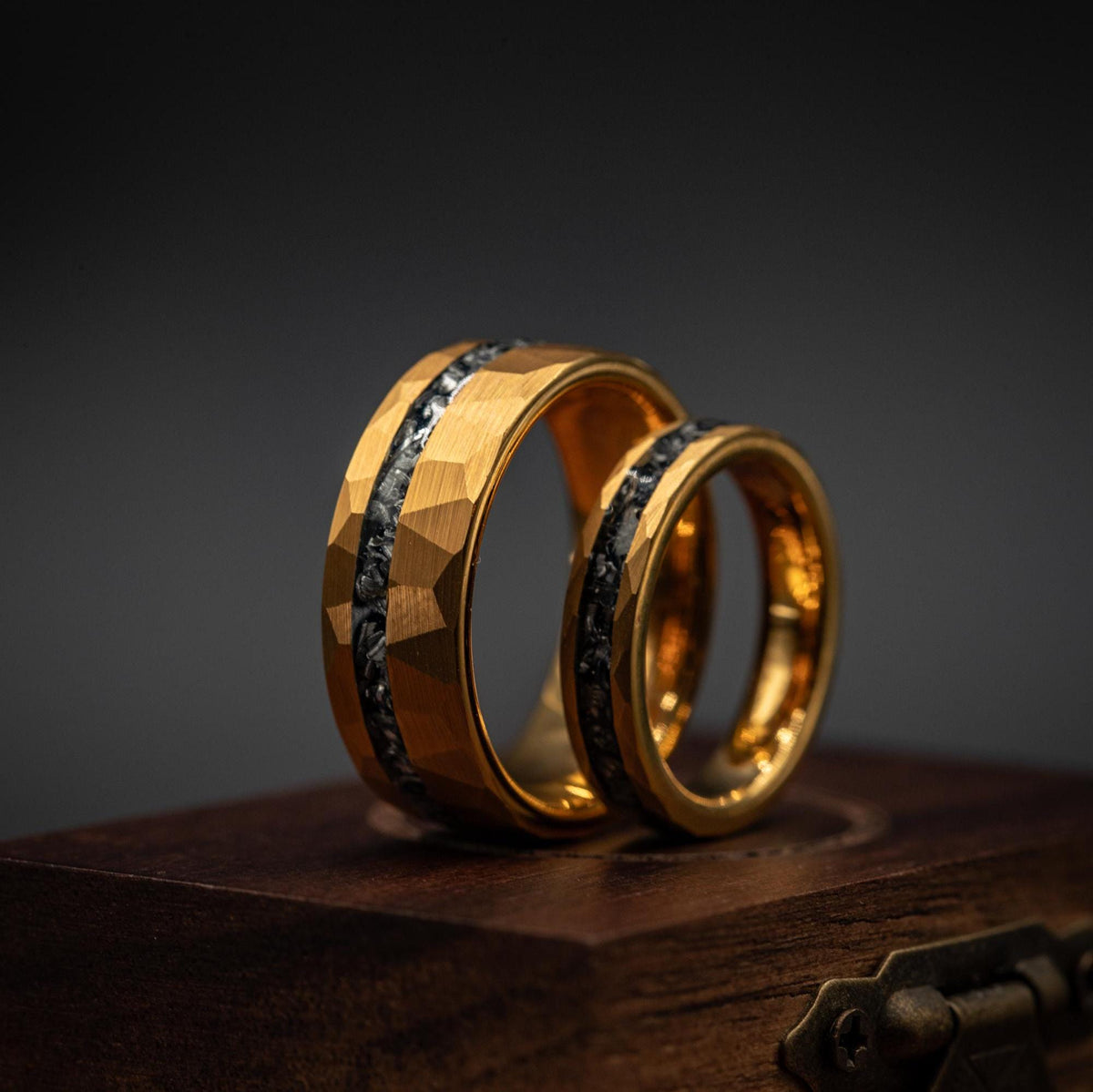 Two Couples Hammered Gold & Meteorite Wedding Bands rest upright on a dark wooden box, showcased against a deep gradient background. The eye-catching design, featuring yellow gold with meteorite-like textured inlays, creates a unique and modern wedding ring set ideal for couples seeking contemporary elegance.