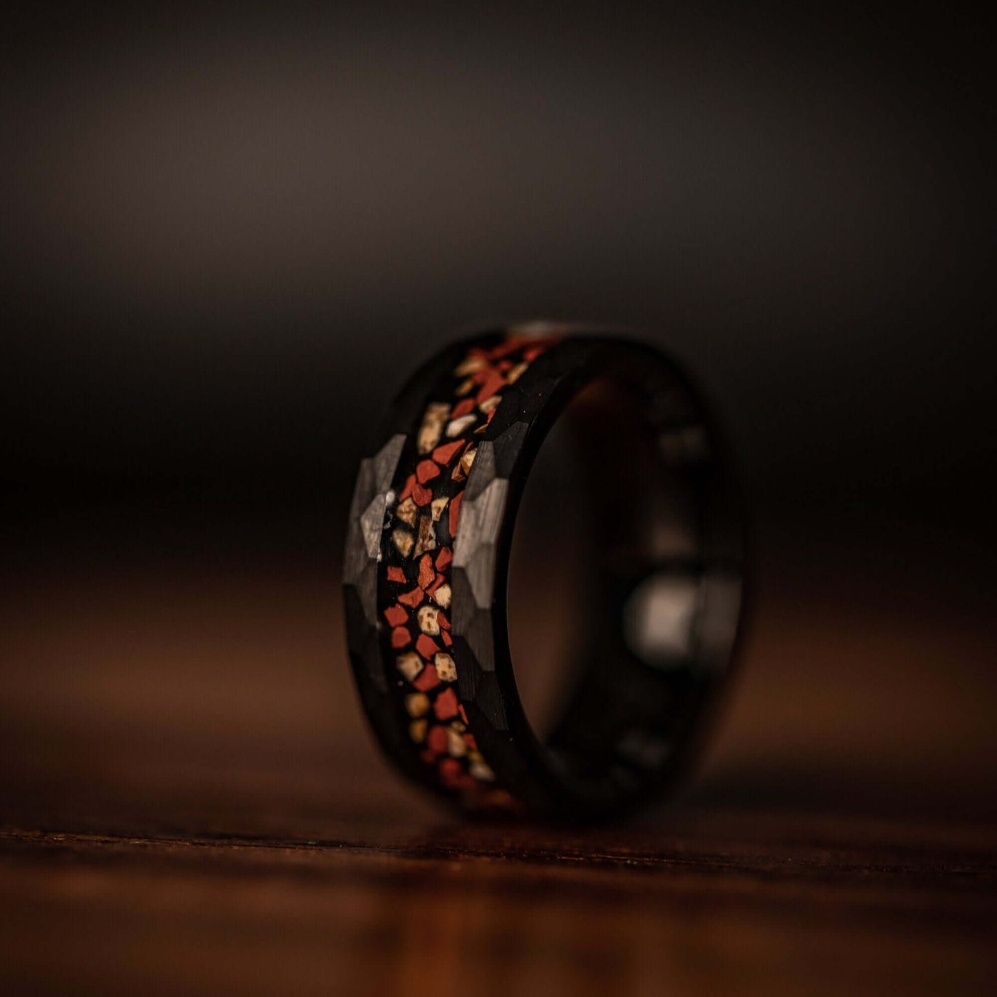 Hammered Black Tungsten Wedding Ring with Red and White Dinosaur Bone Inlay standing on its side on a wooden table